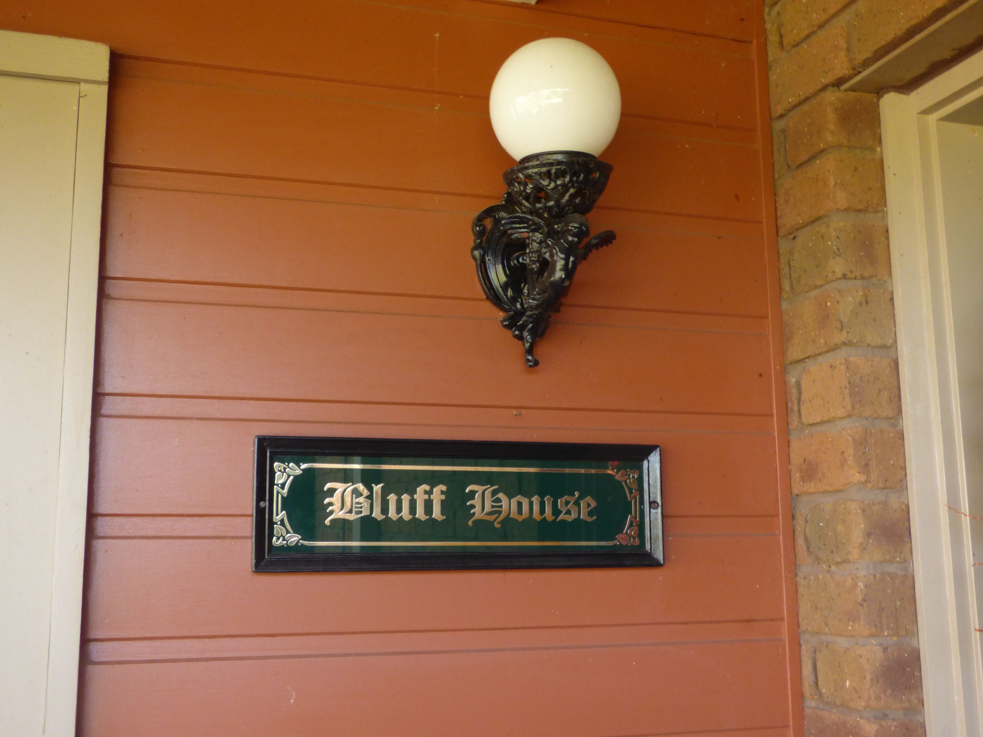 Uma placa com a inscrição 'Bluff House' fixada numa parede com linhas horizontais e um candeeiro decorativo acima.