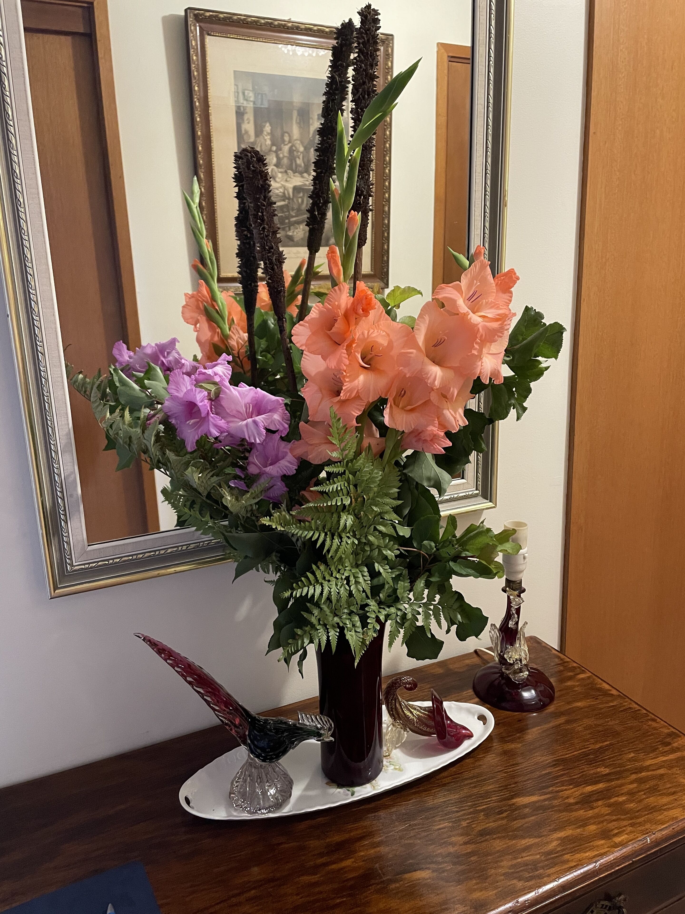 A floral arrangement in a vase on a wooden table with a mirror and framed picture in the background.