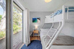 Quarto com beliche e vista para a praia através de uma janela, uma pequena mesa de cabeceira com abajur, e um tapete azul.
