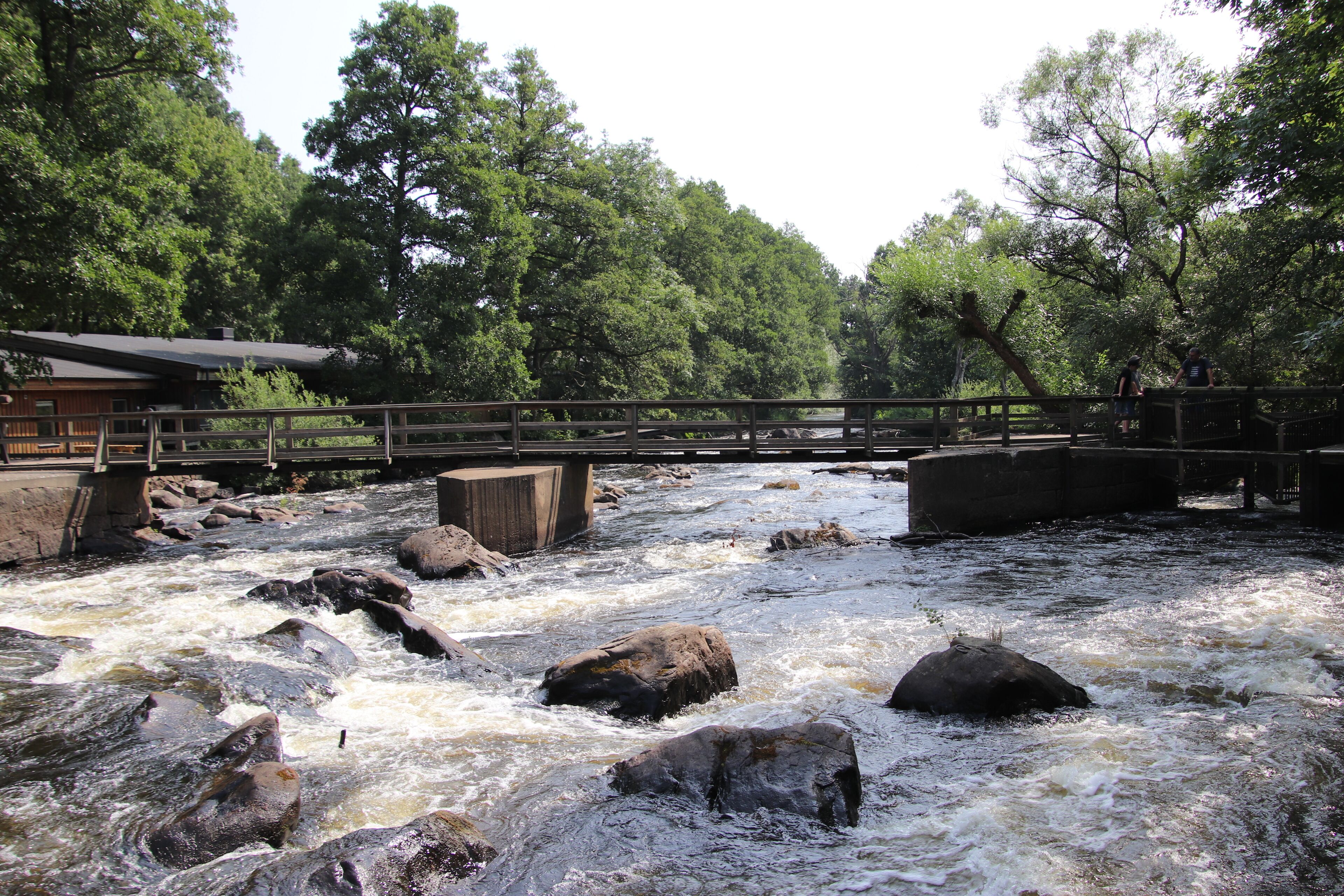 A wooden bridge over a fast-flowing river with rocks, surrounded by dense trees and a building with a sloped roof.