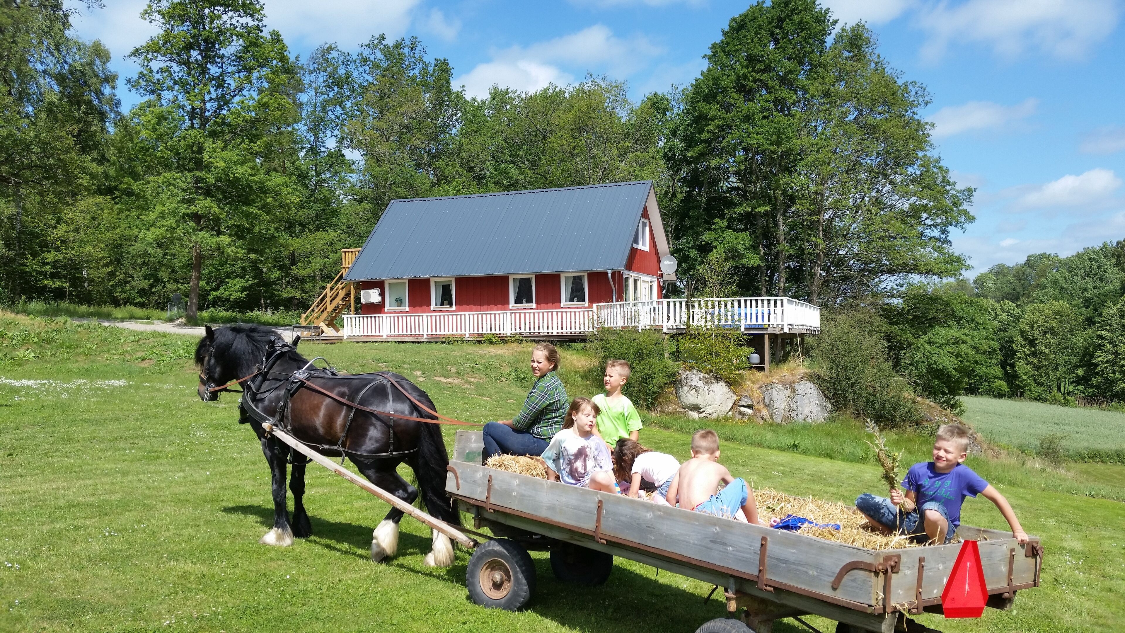 Un groupe de personnes est assis dans une charrette en bois tirée par un cheval, avec une maison rouge en arrière-plan.