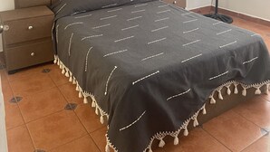 Une chambre à coucher avec une tête de lit capitonnée, un lit recouvert d’une couvre-lit sombre, une table de chevet, une commode, un ventilateur sur pied et un ventilateur de plafond.