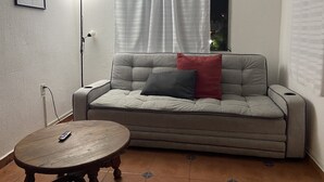 Un salon avec un canapé, une table ronde en bois, une lampe et une fenêtre avec des rideaux.