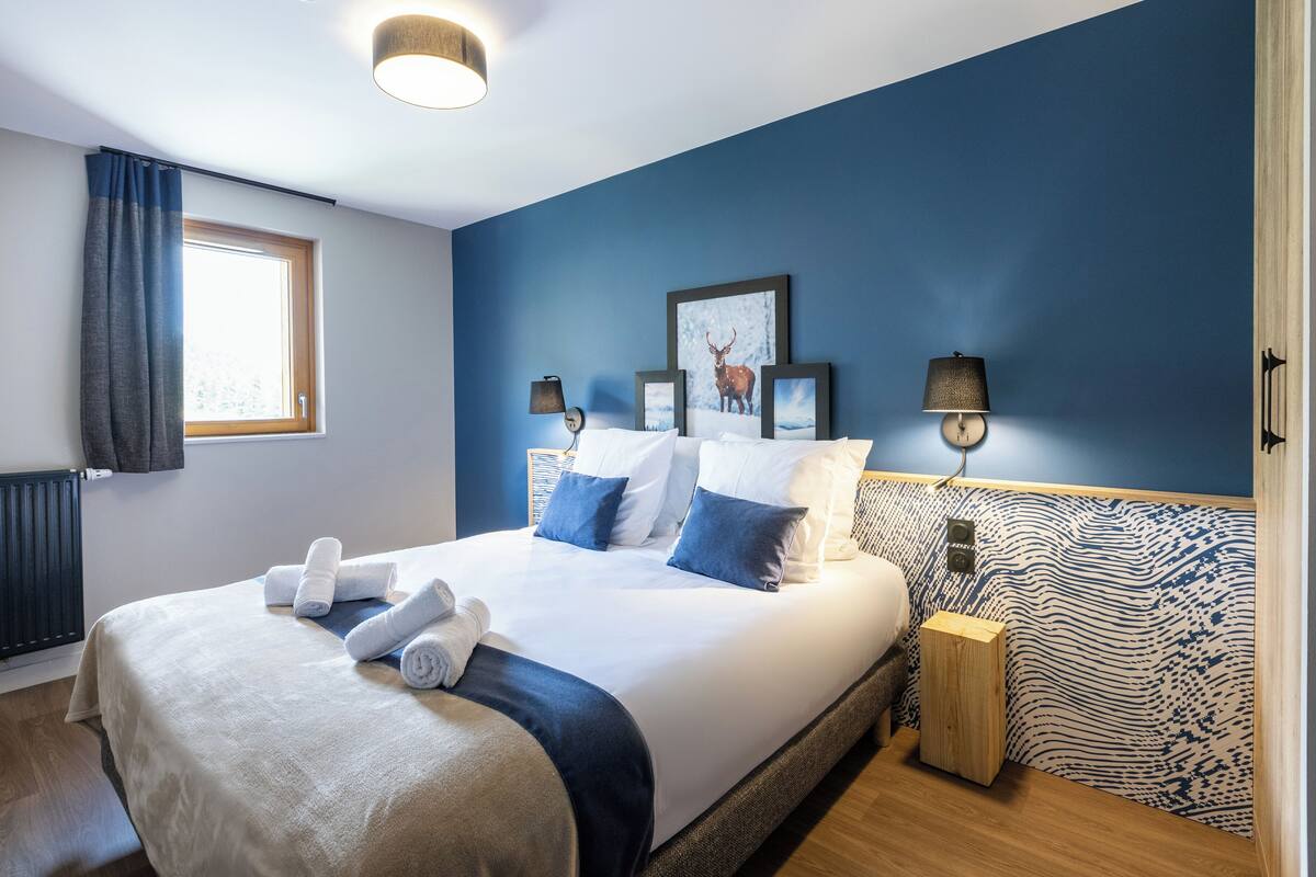 A hotel room with a large bed, two wall-mounted lamps, a window with curtains, and a radiator.