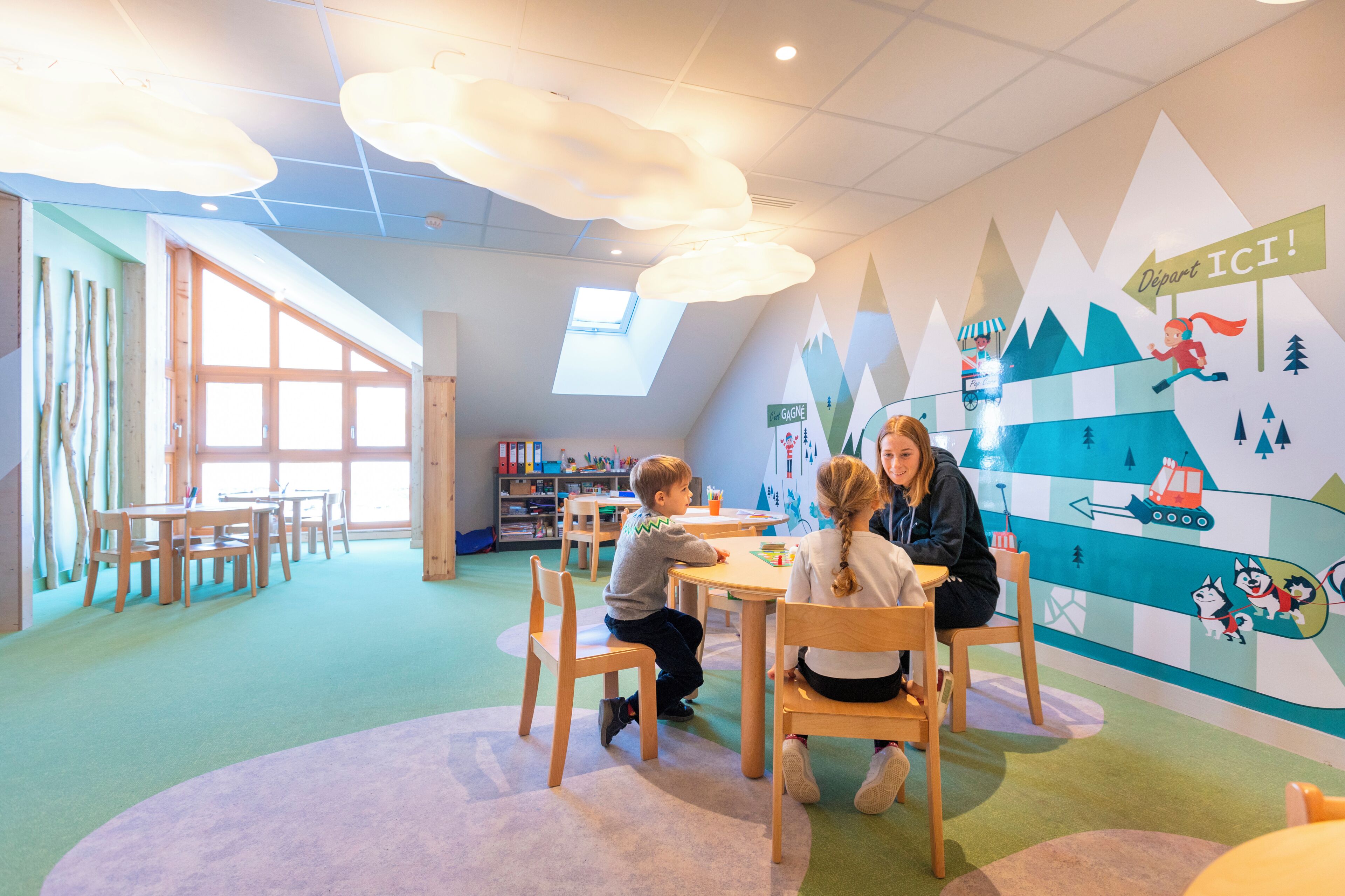 A children's play area with tables and chairs, a large window, and a wall mural with a train and mountains.