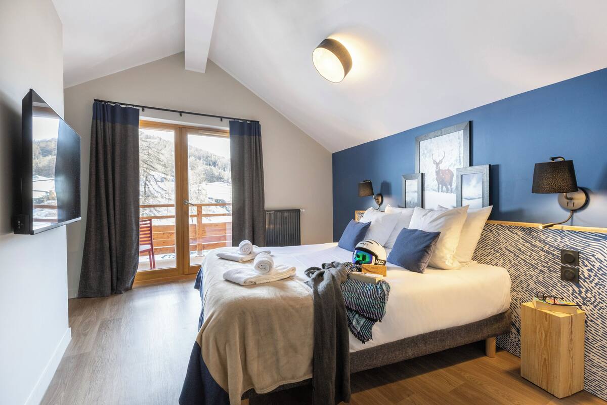 A modern hotel room with a large bed, a television, a balcony with a view of snow-covered hills, and a blue accent wall.