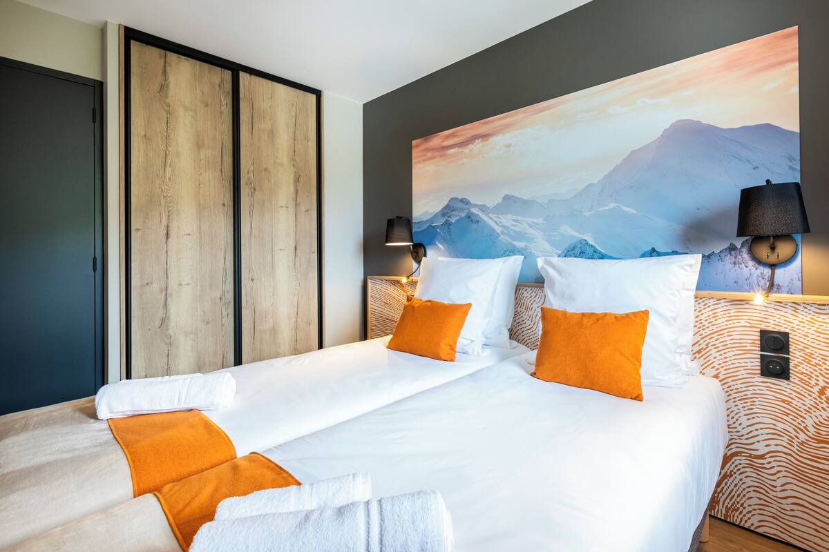 A hotel room with a large bed, two bedside tables, a wooden wardrobe, and a mountain landscape painting on the wall.