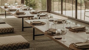 Uma sala de jantar com uma mesa comprida posta para refeição, grandes janelas com vista para o exterior e decoração rústica.