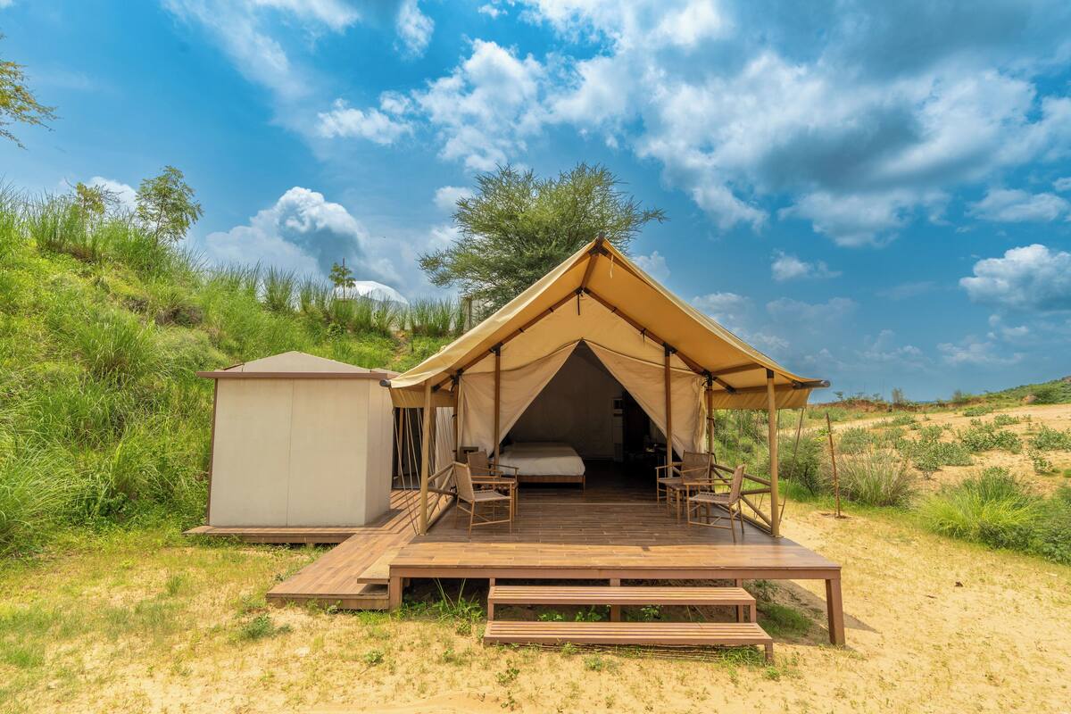 A safari tent with a wooden deck, a small attached structure, and a view of the surrounding landscape.