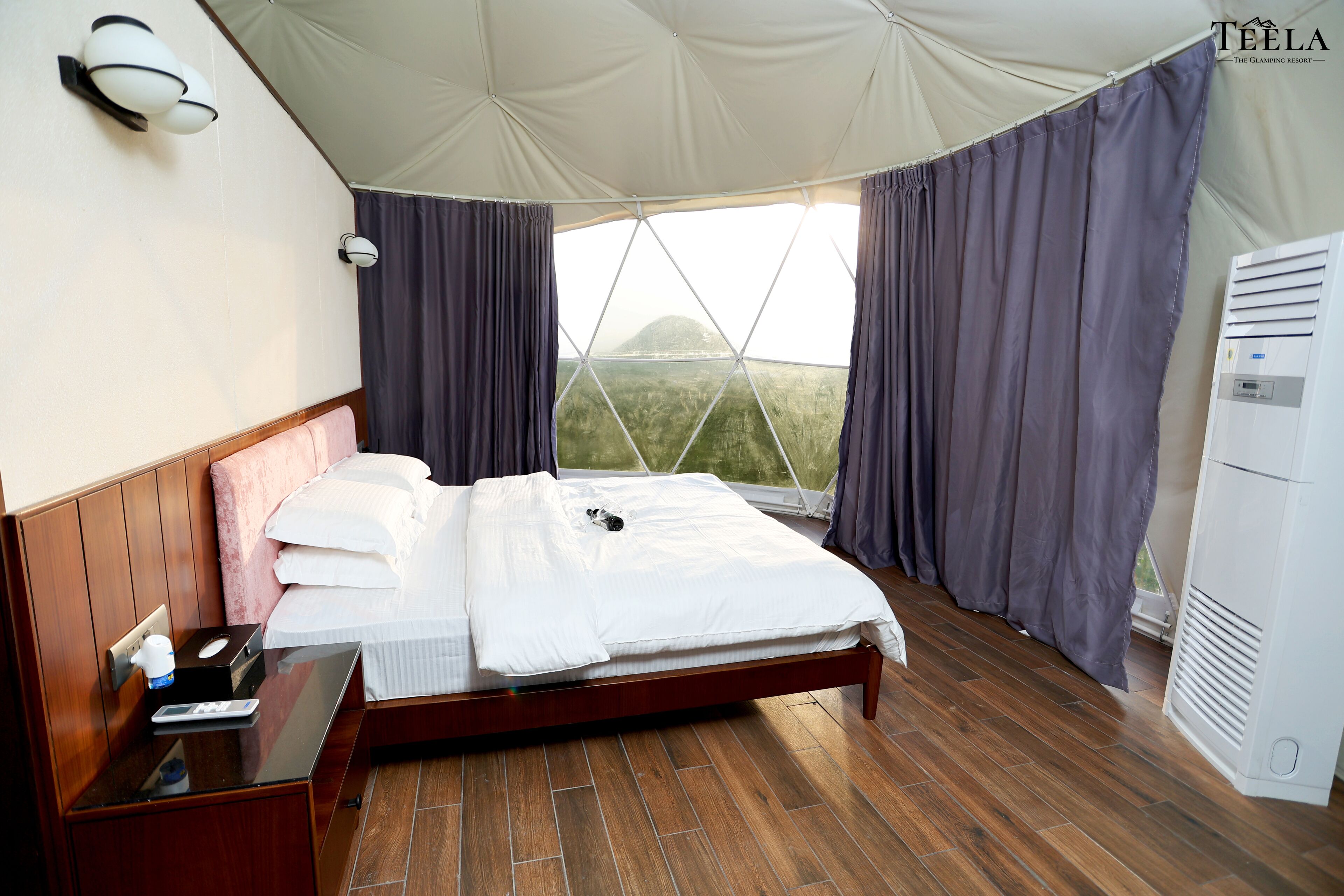 A bedroom with a wooden bed, a bedside table, a window with curtains, and a wall-mounted light fixture.