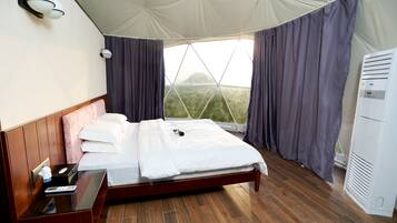 A bedroom with a wooden bed, a bedside table, a window with curtains, and a wall-mounted light fixture.