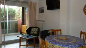 Ein Essbereich mit einem runden Tisch, der mit einer blau-gelben Tischdecke bedeckt ist, zwei Holzstühlen, einem kleinen Tisch mit einer Fernbedienung und einem Fenster mit Jalousien und Vorhängen.