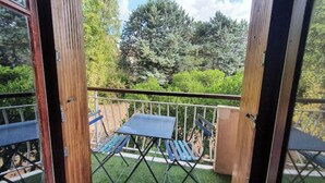 Un balcón con mesa y sillas, rodeado por barandas de madera y vista a árboles y follaje.