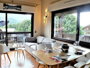 A modern living room with a sofa, dining table, and chairs. There are large windows with a view of greenery and buildings. - Duplex with balcony and parking in Samoëns - Ideal for remote work (Samoëns)