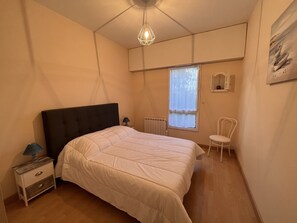A bedroom with a bed, bedside table, chair, window, radiator, and a hanging light fixture. - Beachside Getaway: 2-Bedroom Apartment with Garage, Close to Port & Shops (Les Sables-d'Olonne)