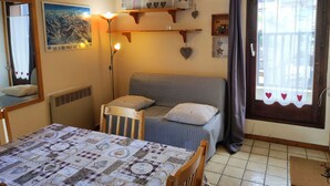 A cozy room with a bed, a table with a quilt, a lamp, and a window with curtains. - Cozy Studio for 4 in Village Center, Near Slopes and Shops (Les Carroz d'Arâches)