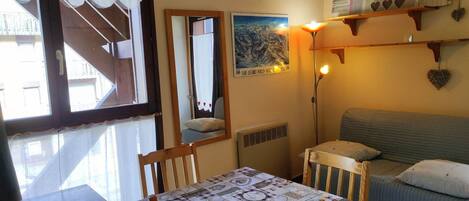 Una habitación acogedora con una mesa de comedor, sillas, un sofá, un espejo y una pared con un cartel y una estantería con adornos.