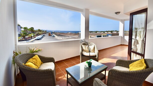 Een balkon met rieten meubilair, een glazen tafel en uitzicht op zee en een parkeerplaats.