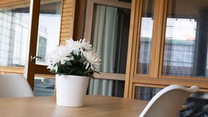 Une table en bois sur laquelle est posé un vase de fleurs blanches, une chaise blanche, et une vue sur un bâtiment à travers une fenêtre.