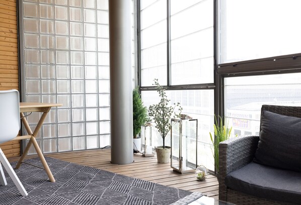 A modern living room with a large window, a grey sofa, a small table, and potted plants.