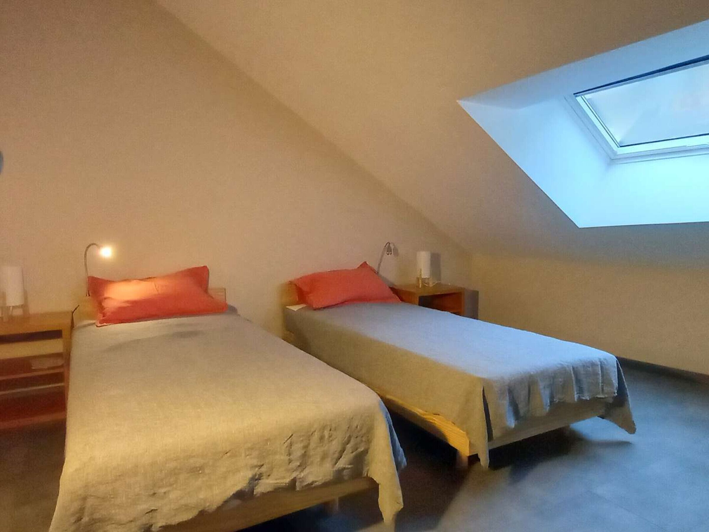 Una habitació petita i poc il·luminada amb dos llits, cadascun amb un coixí i una taula de nit. Es veu un finestral al sostre inclinat.