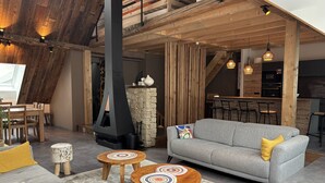 Ein moderner Loft-Wohnraum mit hohen Decken, freiliegenden Holzbalken, einem Steinkamin und einer gemütlichen Sitzecke mit Sofa und runden Tischen.