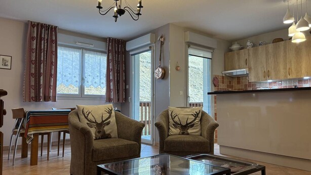 A living room with two armchairs, a glass coffee table, a chandelier, a dining area with a table and chairs, and a kitchen with wooden cabinets and a window. - Cozy Apartment with Balcony, WiFi, and Free Parking near Ski Slopes (Villard-de-Lans)