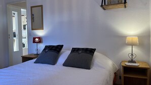 A bedroom with a bed, two pillows, a bedside table with a lamp, a mirror, and a shelf with decorative items. - Cozy Apartment with Balcony, WiFi, and Free Parking near Ski Slopes (Villard-de-Lans)