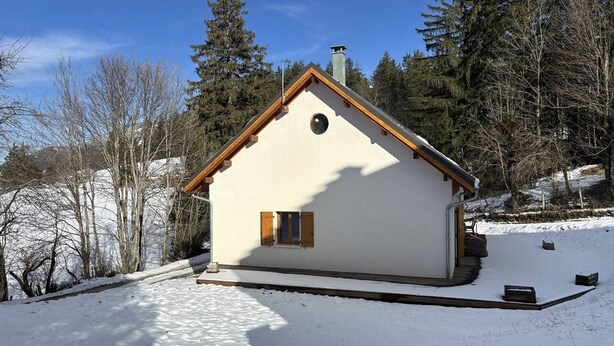 A small, single-story house with a sloped roof, surrounded by snow and trees.