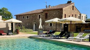 Una piscina cristalina con sillas de descanso y sombrillas, un edificio de piedra con tejado de baldosas y un cielo azul despejado.