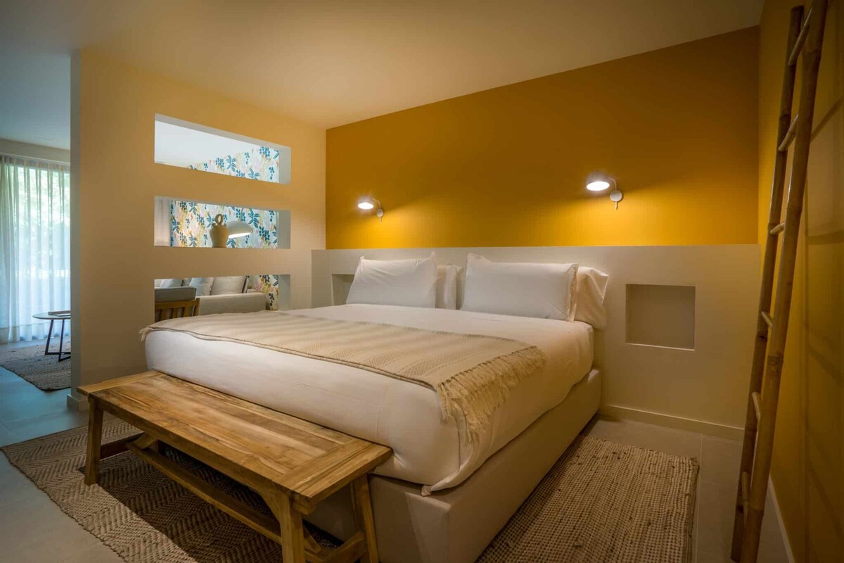 A bedroom with a large bed, a wooden bench, a small table, and a wall-mounted light fixture.