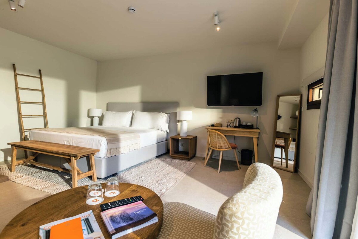 A hotel room with a bed, desk, chair, TV, and a small table with remote controls and books.