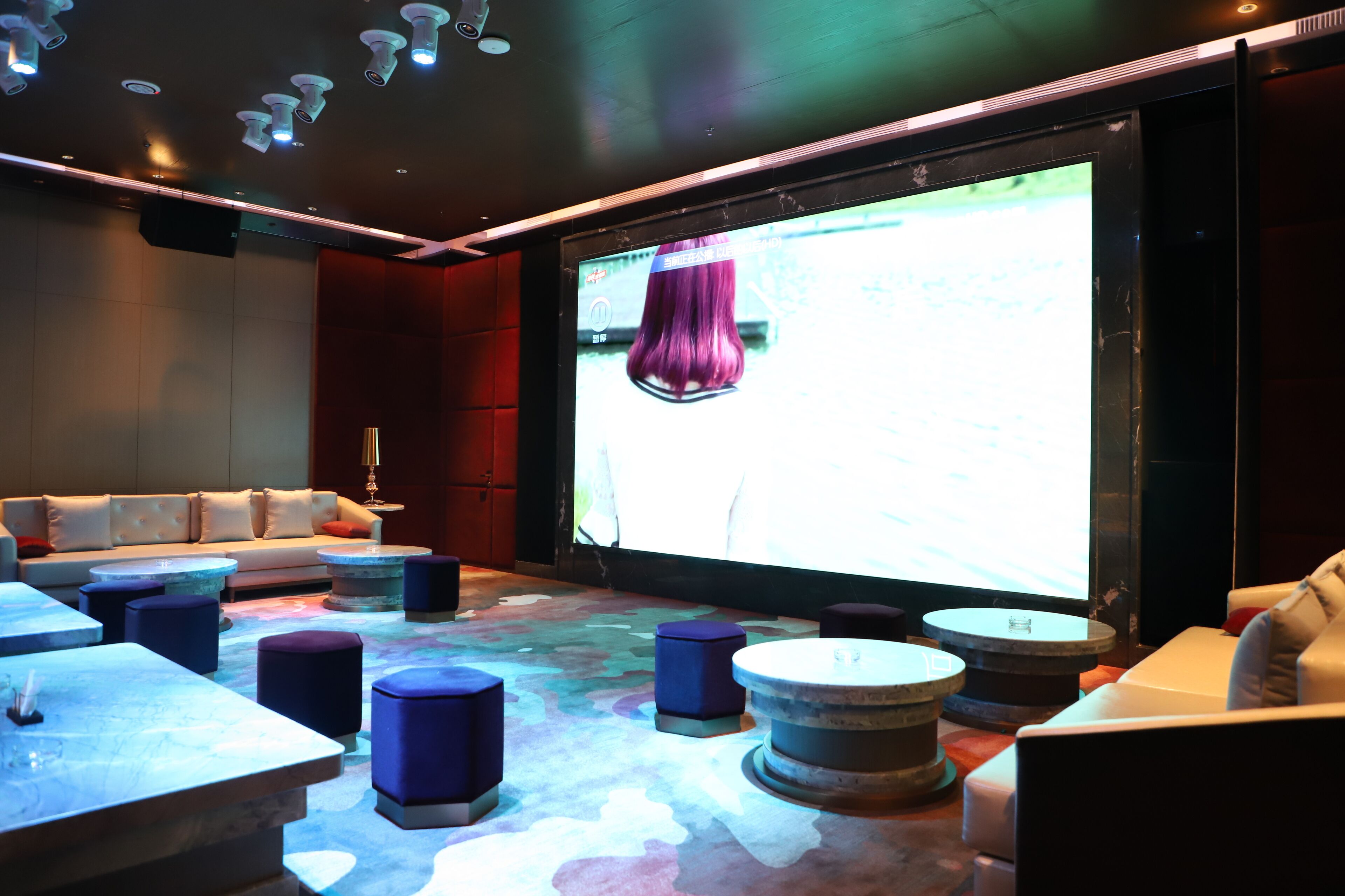 Un espace lounge moderne doté d’un grand écran diffusant une image d’une perruque violette, de fauteuils confortables et de tables rondes originales.