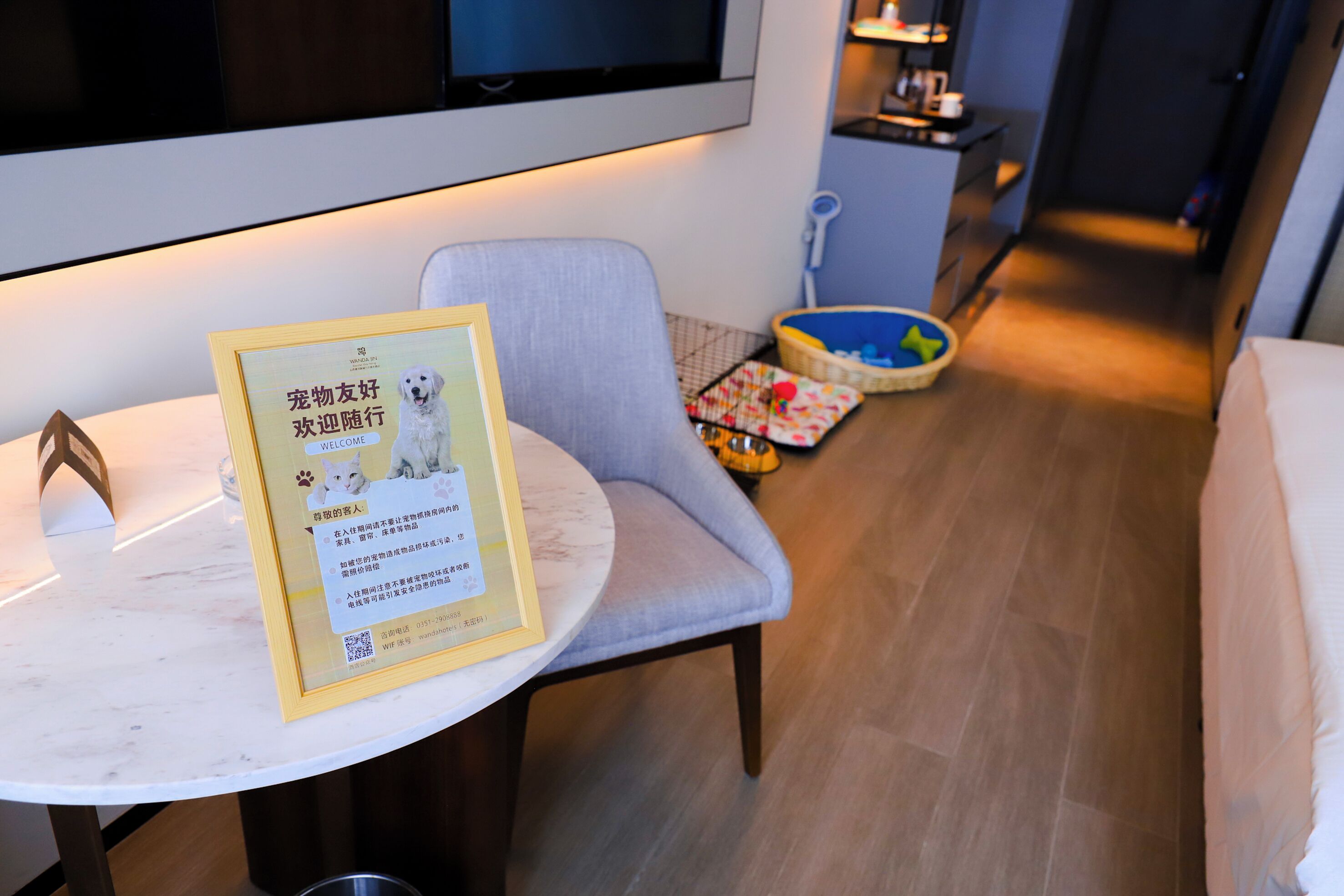 A hotel room with a round marble table, a framed welcome sign, a grey armchair, a pet bed, and a small kitchenette in the background.