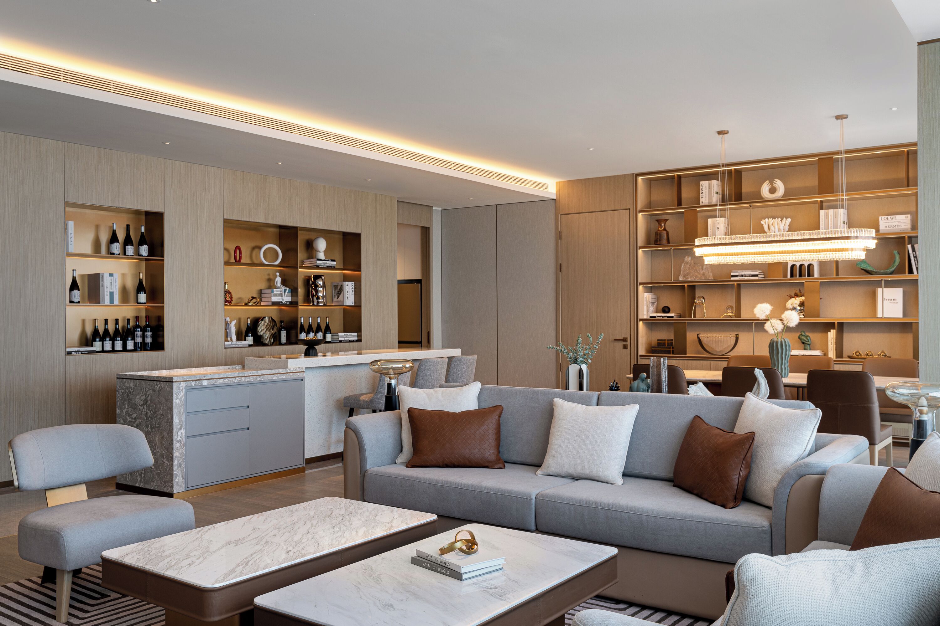 A modern living room with a grey sofa, a marble coffee table, and a kitchen area with wine bottles.