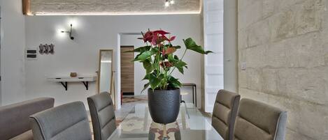 A dining area with a glass table, grey upholstered chairs, a potted plant, and a modern pendant light.