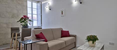 Uma sala de estar moderna com um sofá bege, uma mesa de centro de vidro e uma pequena mesinha lateral com uma planta.