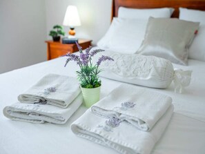 A neatly made bed with white linens and pillows, a small potted plant with purple flowers, and a folded towel with a floral design. - Solar House Babina 1 (Korcula)