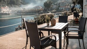 An outdoor patio with wicker chairs and a table, overlooking a body of water and a mountainous landscape. - Solar House Babina 1 (Korcula)