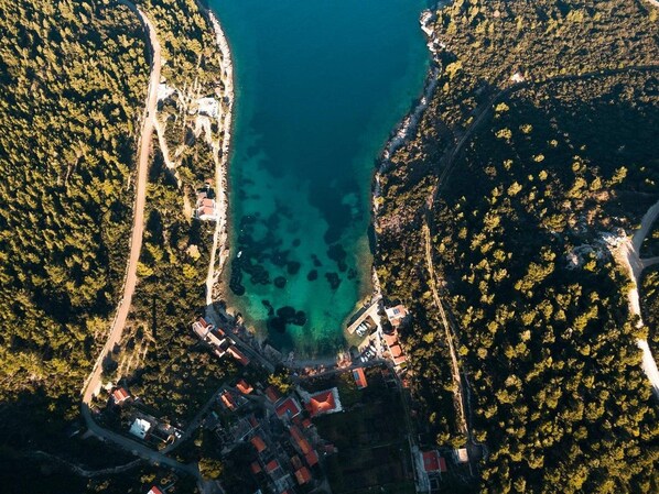 An aerial view of a coastal area with a small settlement, clear turquoise waters, and dense forested hills.