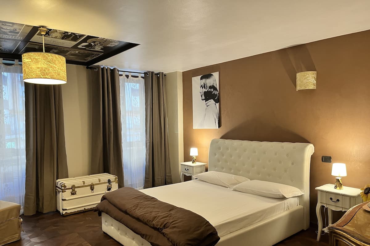 A hotel room with a large bed, two bedside tables with lamps, a chest of drawers, a suitcase, and a large window with curtains.