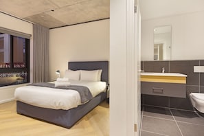 A modern hotel room with a large bed, a bedside table, a mirror, and a bathroom with a sink and toilet. - Bridgewater View 415 by HostAgents  (Cape Town)