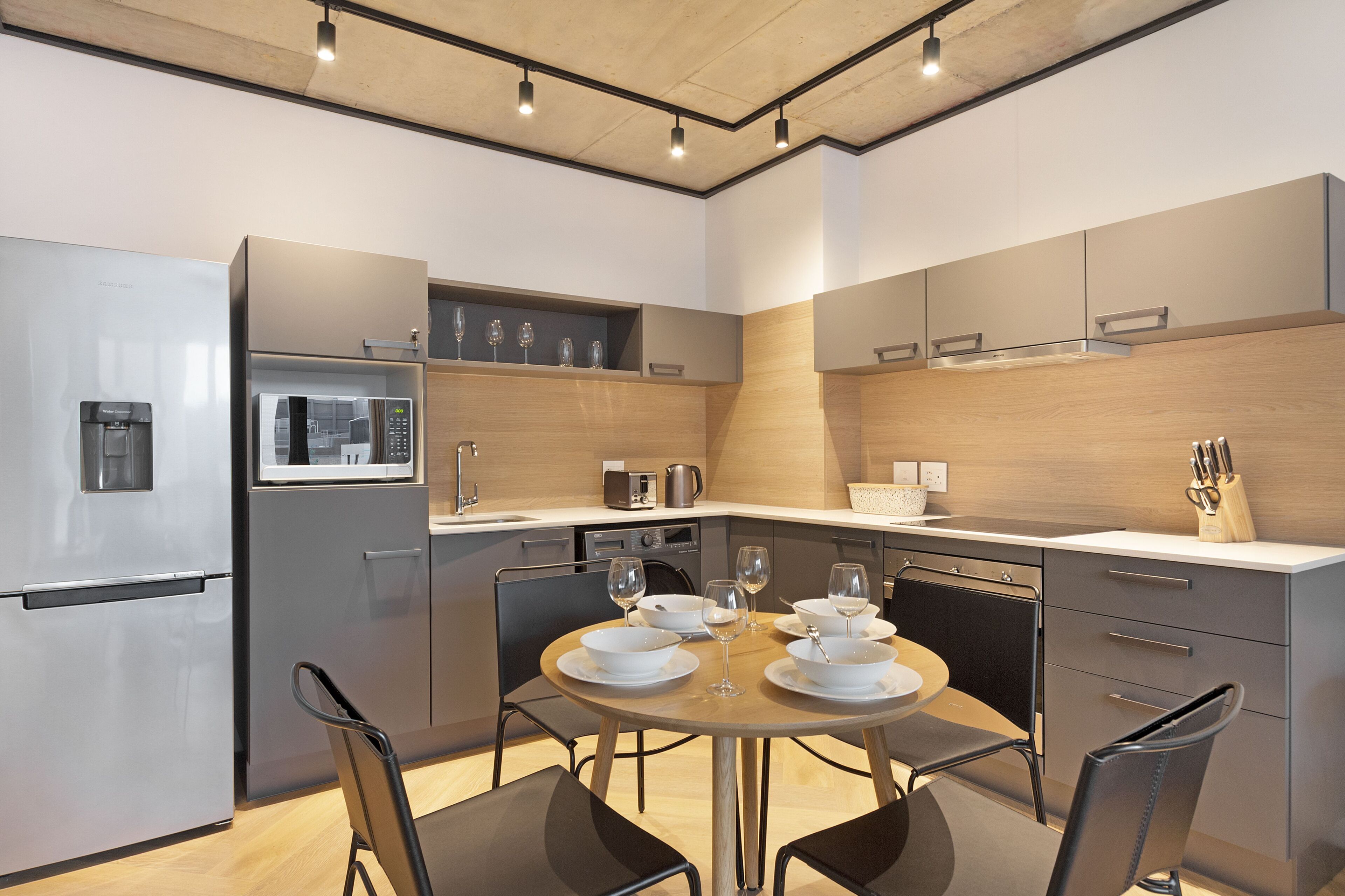 A modern kitchen with a built-in microwave, oven, and coffee maker. There is a dining table with chairs and a set of wine glasses.