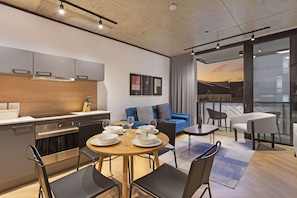 A modern kitchen with stainless steel appliances, a dining area with a round table and chairs, a blue sofa, and a balcony with a view of the sunset. - Bridgewater View 415 by HostAgents  (Cape Town)