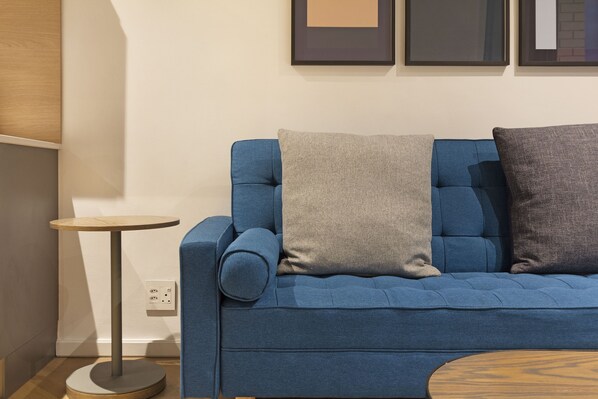 A blue sofa with two cushions, a small round table, and a wall-mounted light switch.