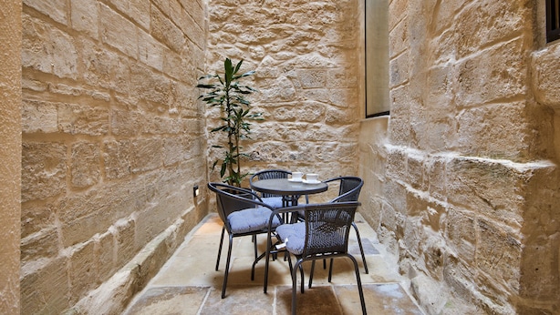 A narrow stone-walled courtyard with a round table and four chairs, a potted plant, and a wall-mounted light fixture. - Vallettastay - Casa Zoe  (Valletta)