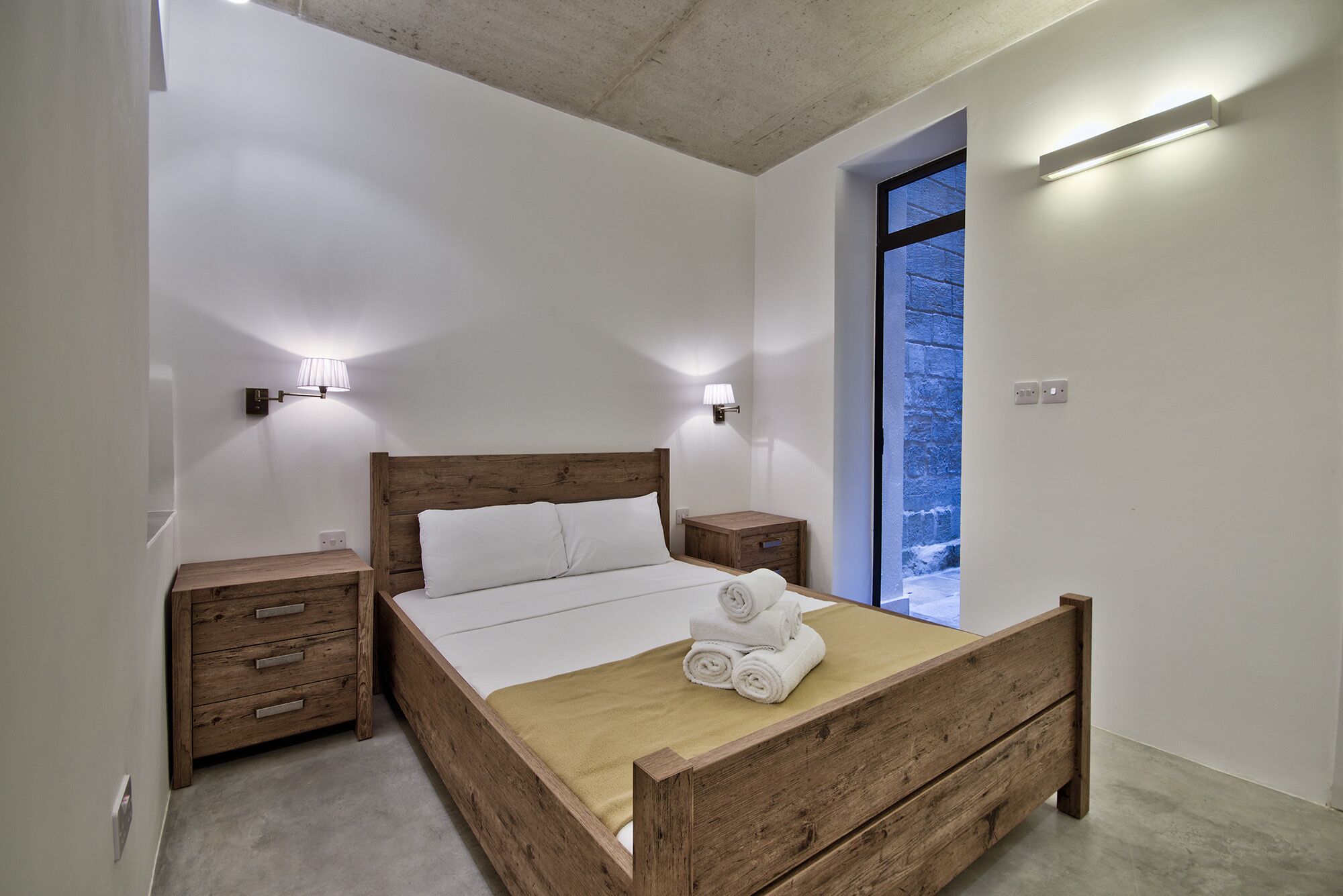 Ein Schlafzimmer mit einem hölzernen Bett, zwei Nachttischen, einem Beistelltisch und einem großen Fenster mit Blick auf einen blauen Himmel.