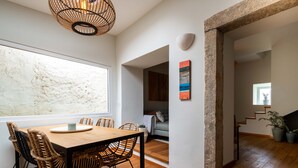 A dining area with a wooden table and wicker chairs, a large window, and a stone wall. - SEA´YA Villa Areia with swimming pool and 5 minutes from the beach (Cascais)