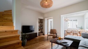 A modern living room with a wooden staircase, a flat-screen TV, wicker furniture, and a large window. - SEA´YA Villa Areia with swimming pool and 5 minutes from the beach (Cascais)