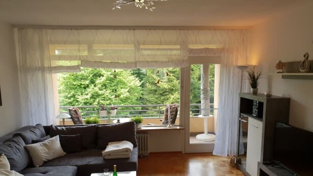 A living room with a sofa, coffee table, and a TV. There is a balcony with plants and a view of greenery.